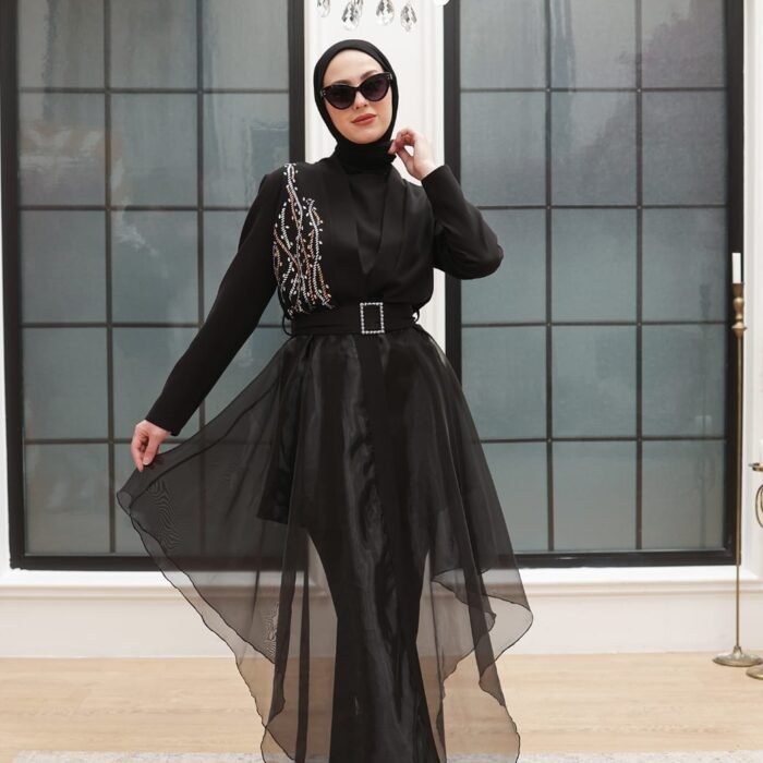 Model wearing a black two-piece evening set with sheer flowing overlay skirt and embellished details, styled with black heels.